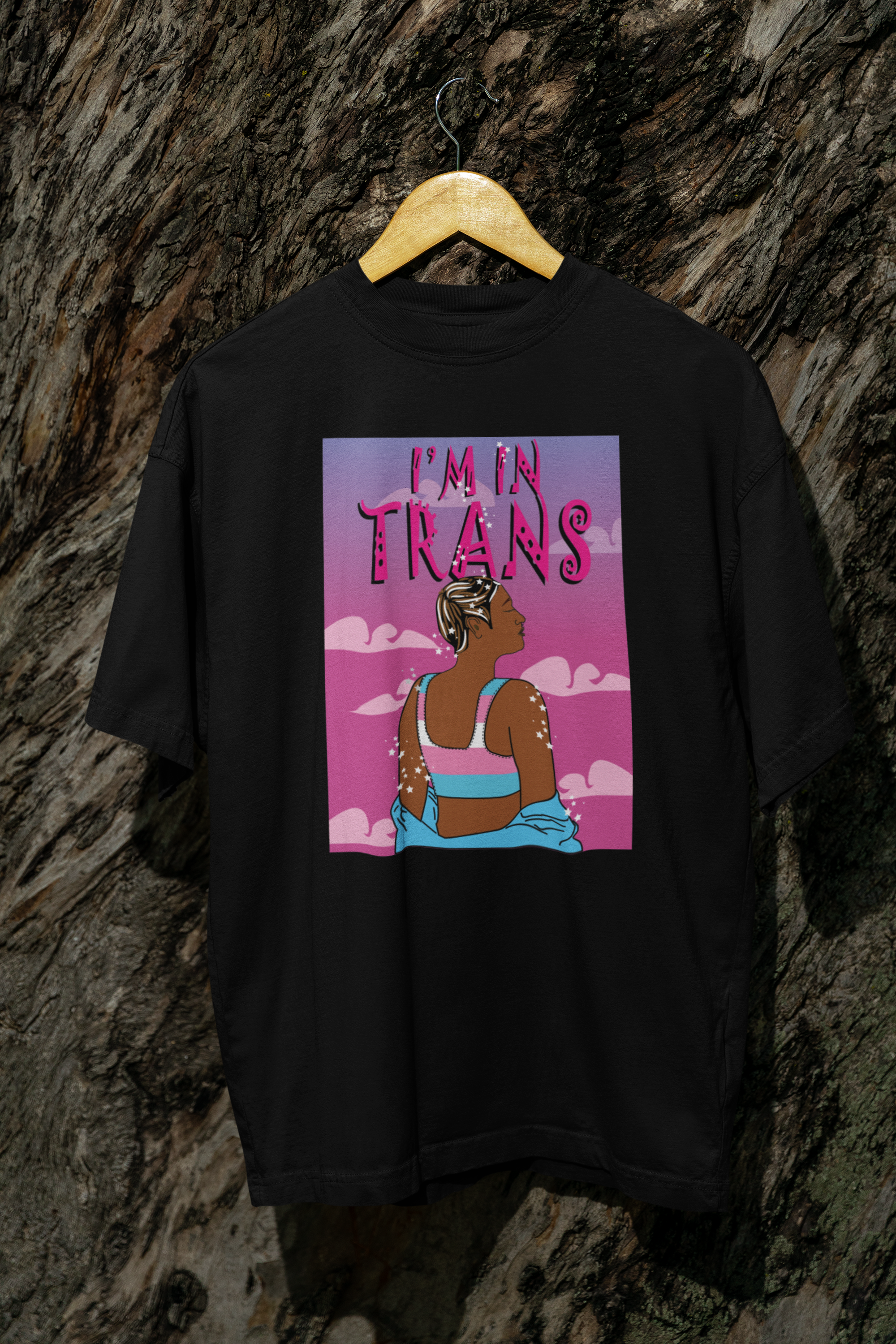 Black unisex t-shirt hanging outdoors against a textured tree bark, showcasing a powerful graphic of a trans person in a trans pride binder surrounded by celestial sparkles and a pink cloudy sky, with the statement “I’M IN TRANS” in bold, stylized font.