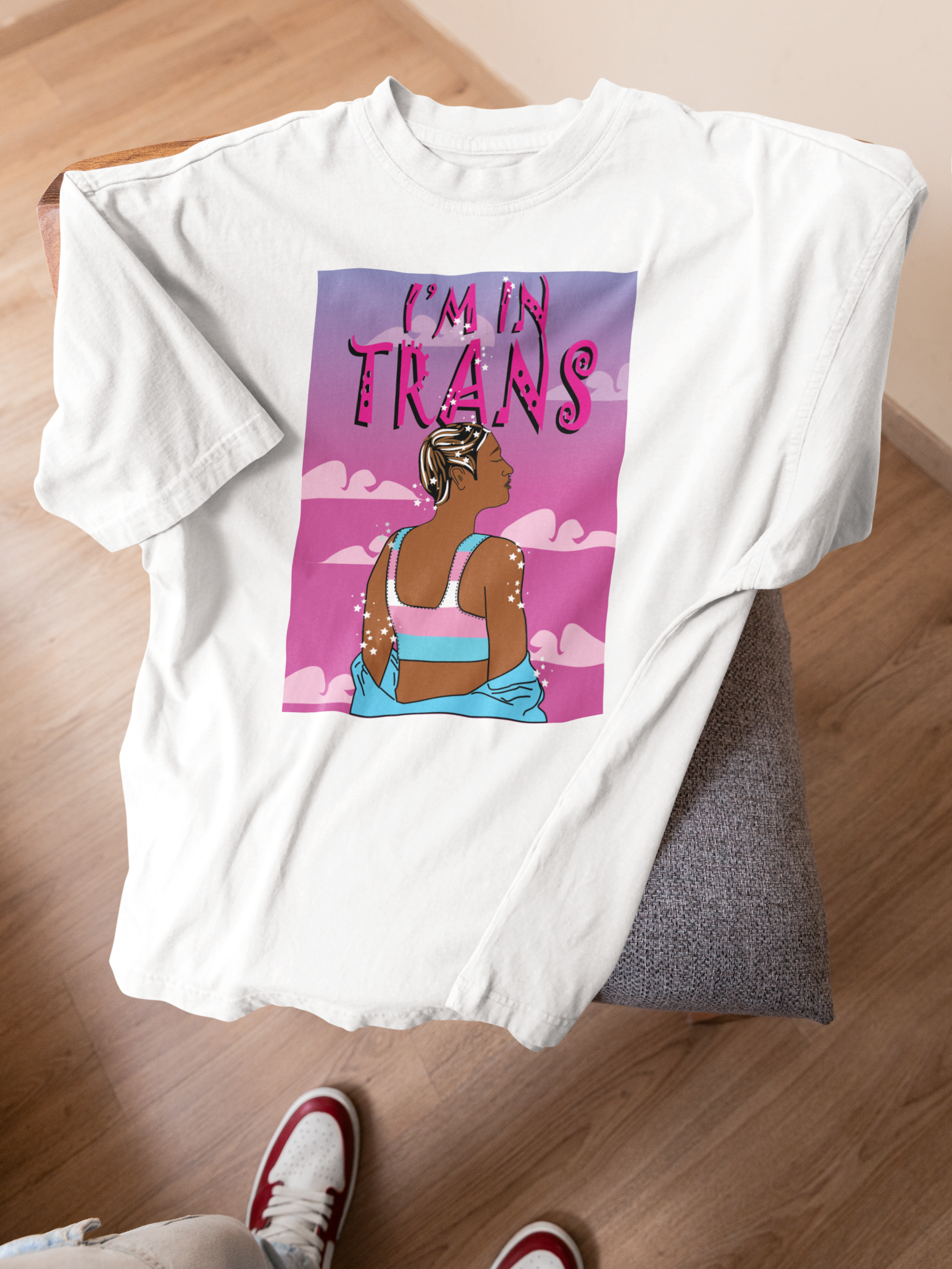 White gender-neutral t-shirt draped on a chair, featuring a bold, illustrated graphic of a trans person from behind wearing a trans pride crop top, set against a dreamy pink and purple cloudscape with the empowering text “I’M IN TRANS” in stylized letters above.