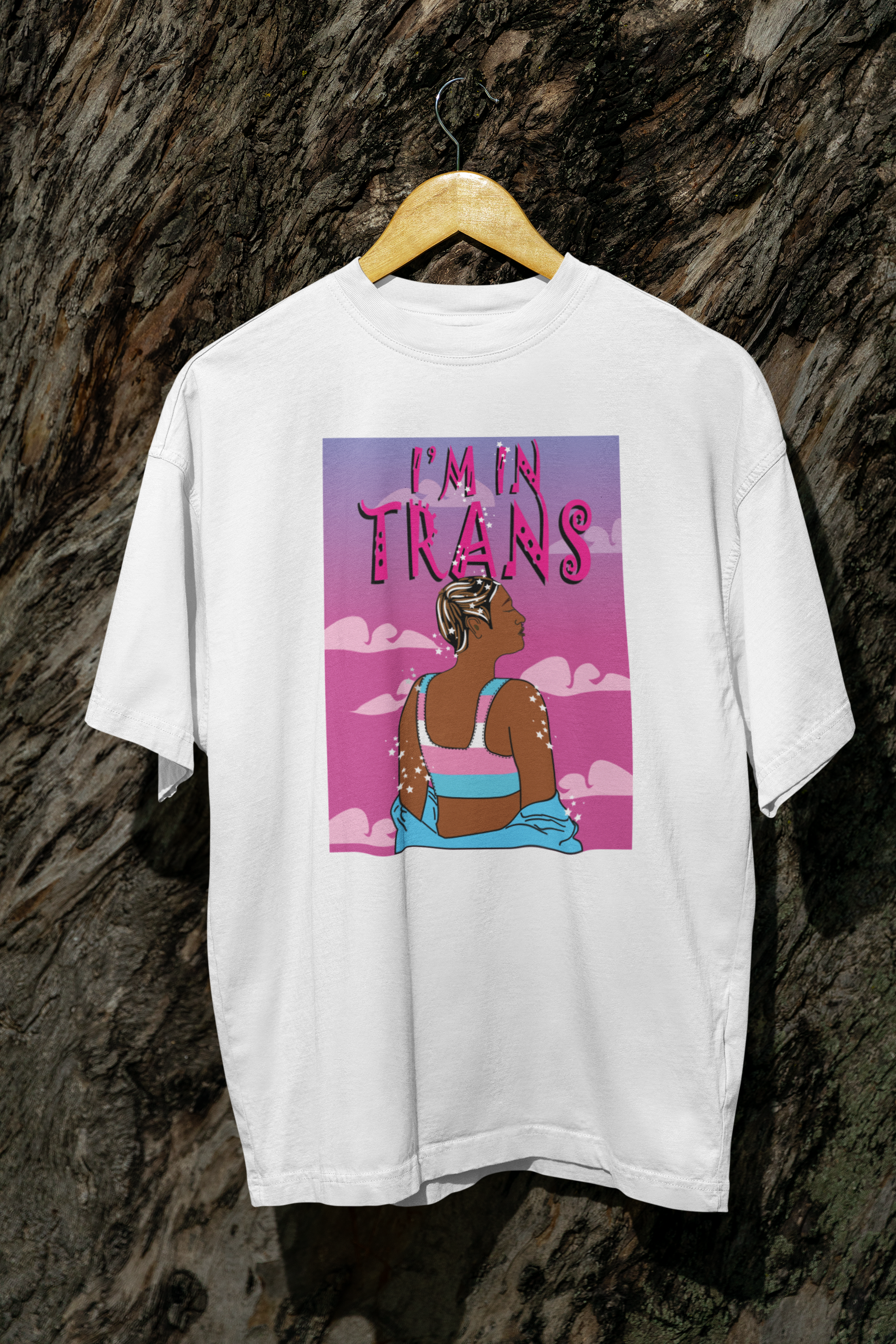 White gender-fluid t-shirt hanging naturally on a tree, displaying a striking illustration of a trans figure in a trans flag crop top, gazing into a dreamy pink sky, with “I’M IN TRANS” boldly printed above in stylized typography.