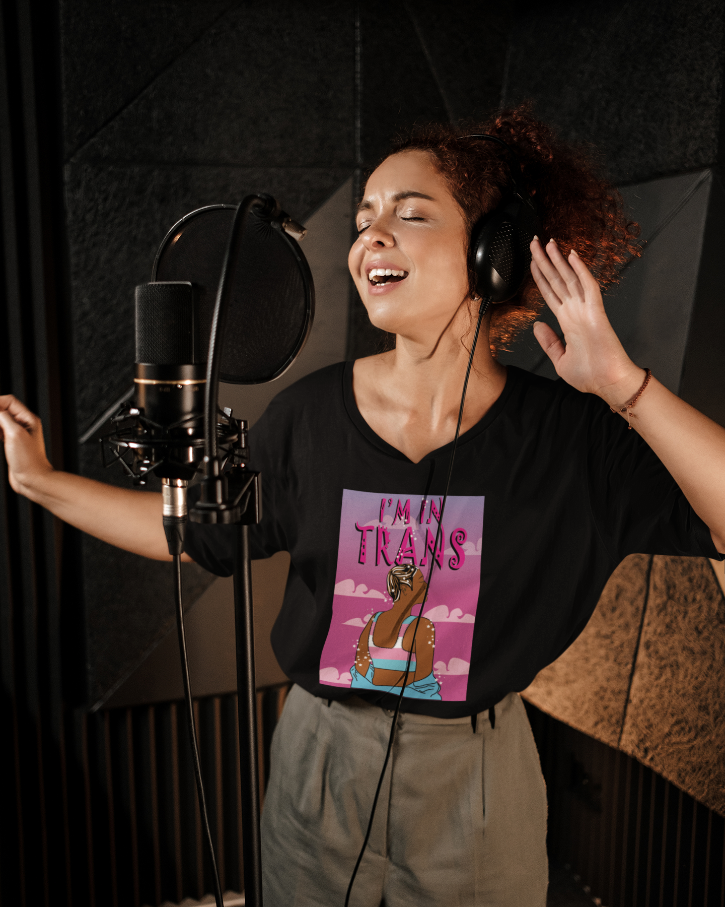 A joyful individual recording in a studio, wearing a black “I’m Trans” graphic T-shirt featuring a striking trans woman against a pink sky backdrop—radiating authenticity and LGBTQIA+ pride.