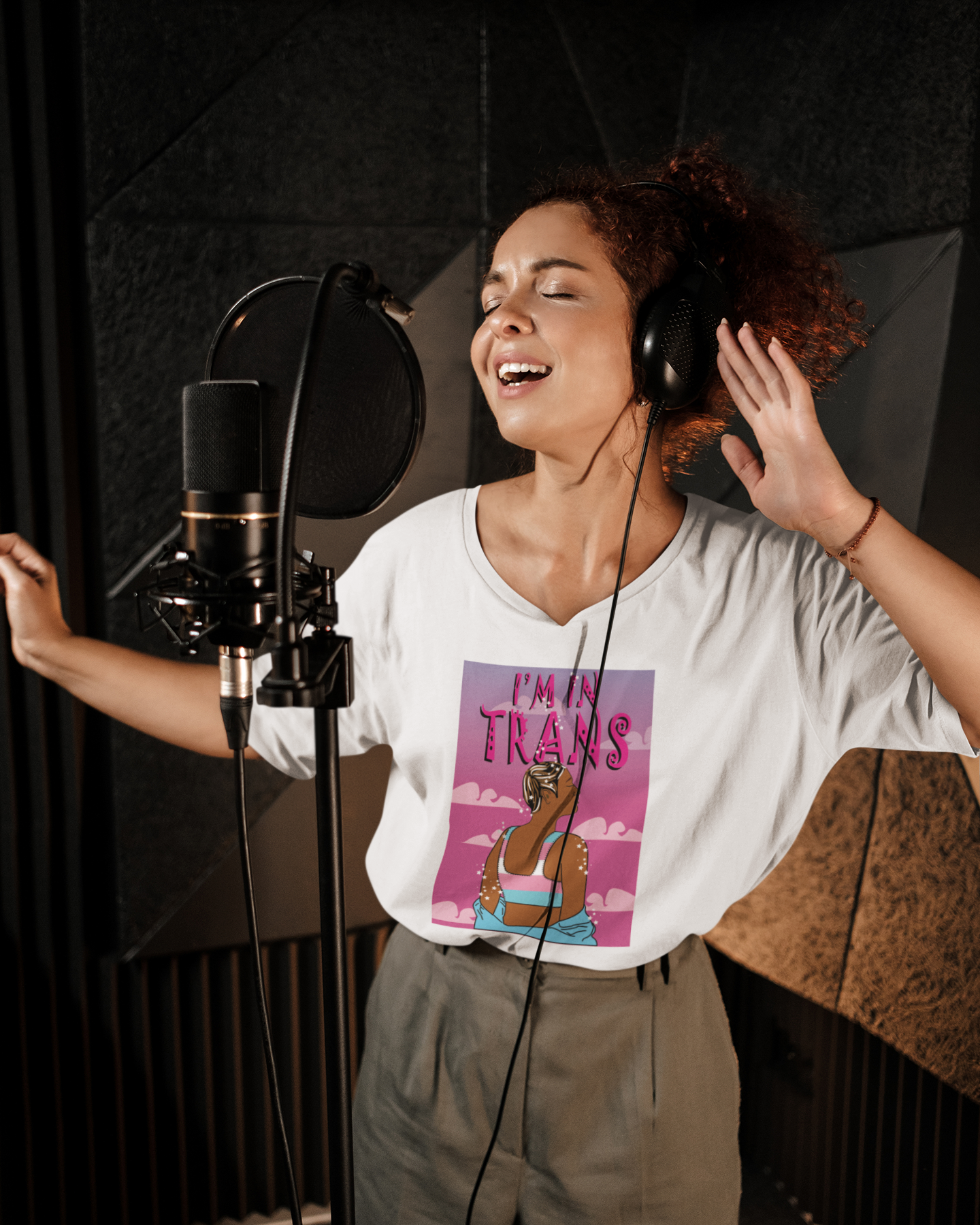 A confident, expressive person wearing a white “I’m Trans” graphic T-shirt with a vibrant illustration of a transgender woman wrapped in the trans flag, singing passionately into a studio mic—boldly celebrating trans identity and self-expression.