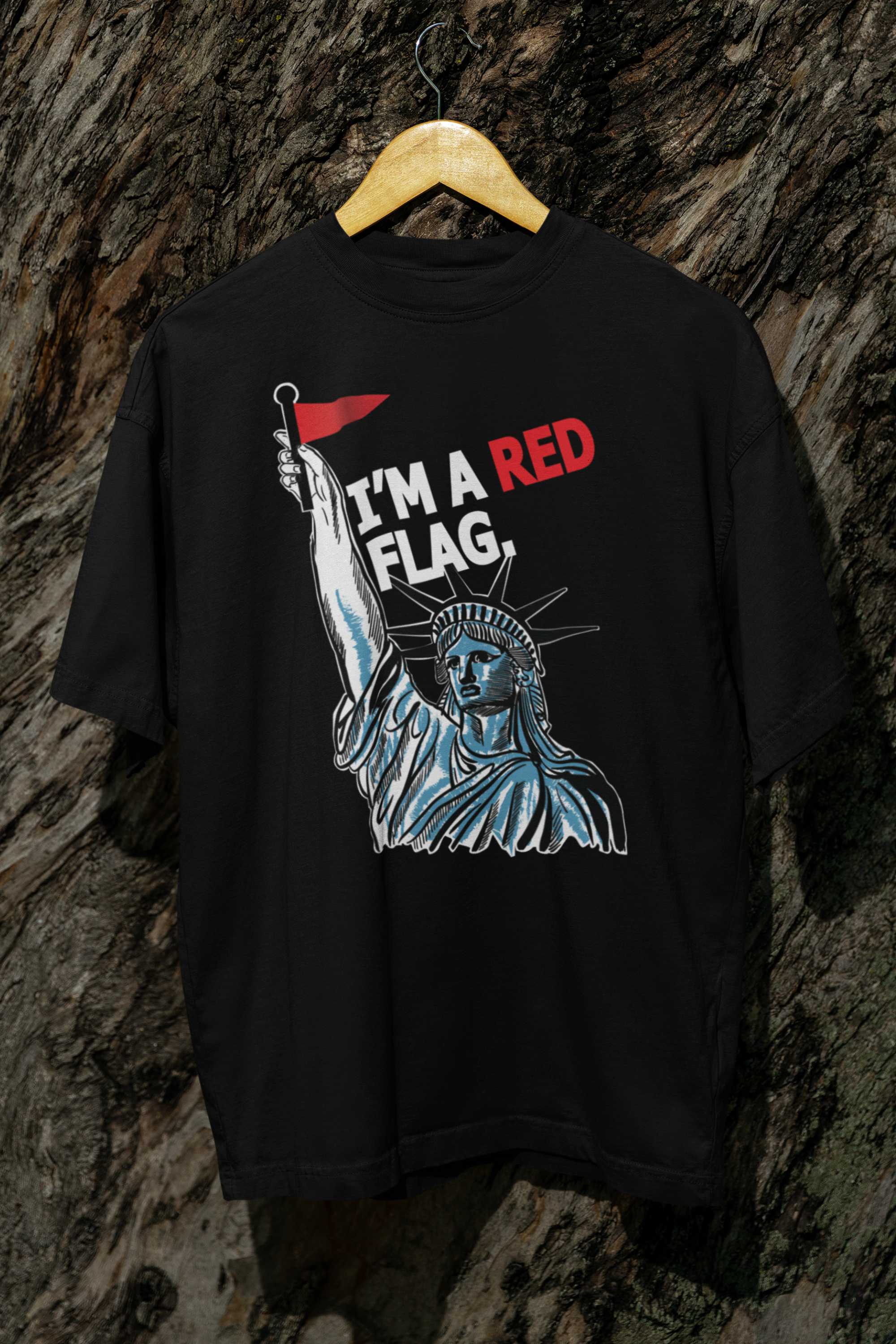 Black unisex t-shirt hanging from a wooden hanger on a rugged tree surface. Bold artwork of the Statue of Liberty holding a red flag, paired with the striking phrase “I’M A RED FLAG” in white and red letters. Designed by ANTI to challenge norms and amplify queer voices through streetwear.

