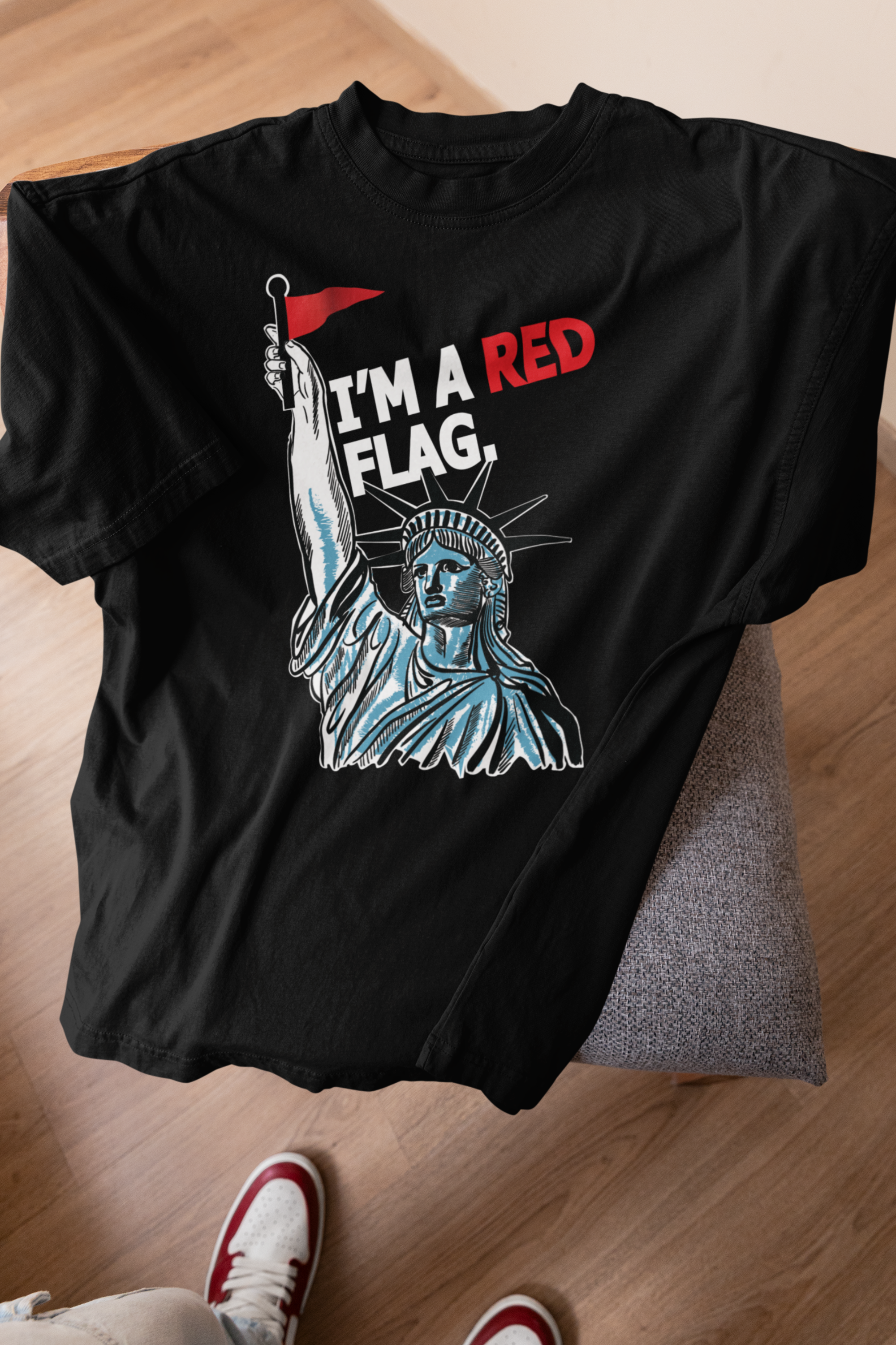 Black t-shirt laid flat on a cushioned chair, with red and white sneakers partially visible below. The shirt features the Statue of Liberty raising a red flag, paired with the bold phrase “I’M A RED FLAG.” A signature piece from ANTI’s LGBTQIA+-inclusive, gender-neutral streetwear collection.

