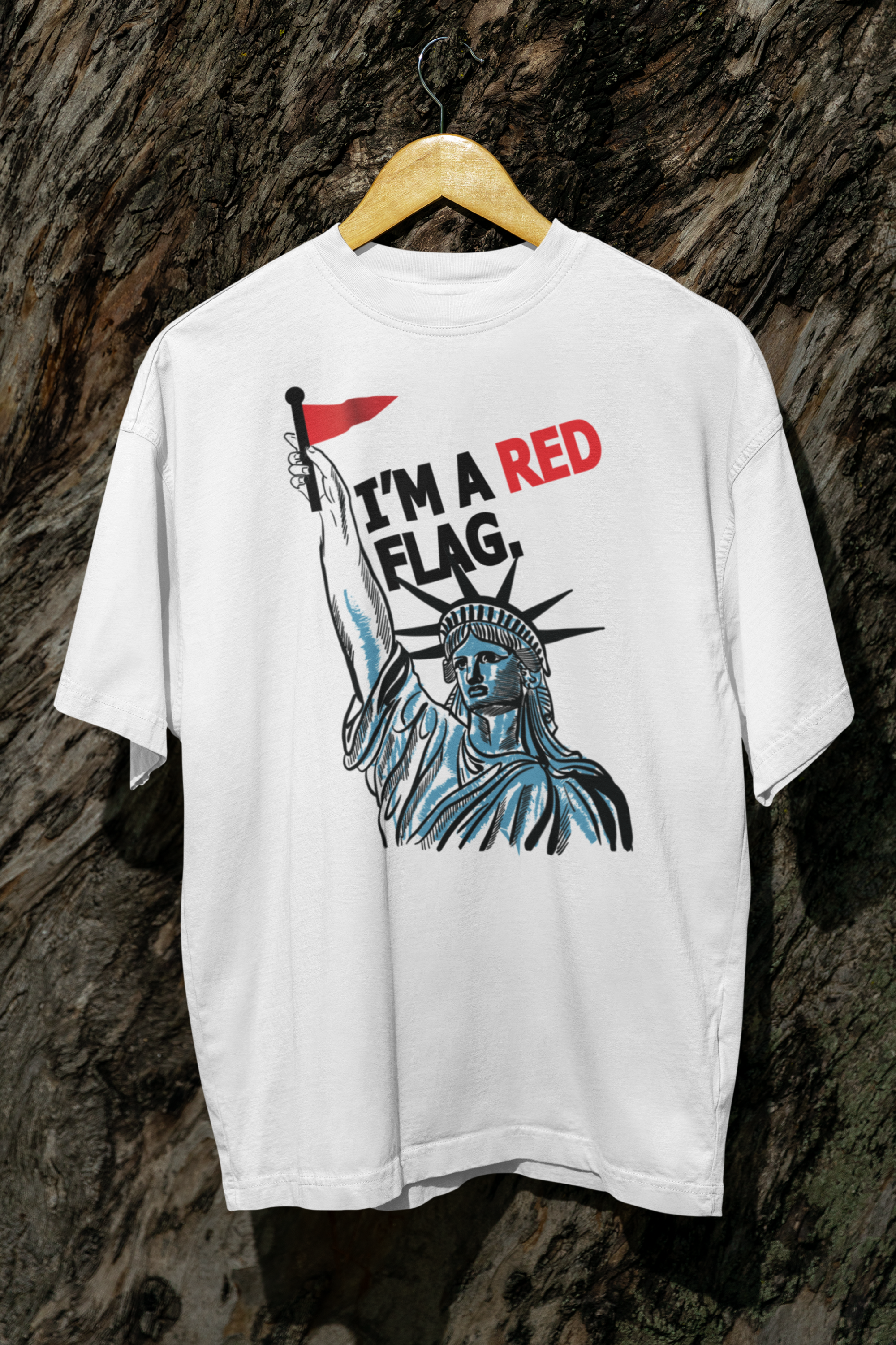White unisex t-shirt from ANTI’s inclusive apparel line hanging on a wooden hanger against a textured tree bark background. The shirt displays a powerful graphic of the Statue of Liberty holding a red flag with bold black and red text reading “I’M A RED FLAG,” designed to challenge societal norms and empower queer, trans, and nonbinary identities.

