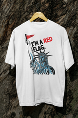 White unisex t-shirt from ANTI’s inclusive apparel line hanging on a wooden hanger against a textured tree bark background. The shirt displays a powerful graphic of the Statue of Liberty holding a red flag with bold black and red text reading “I’M A RED FLAG,” designed to challenge societal norms and empower queer, trans, and nonbinary identities.

