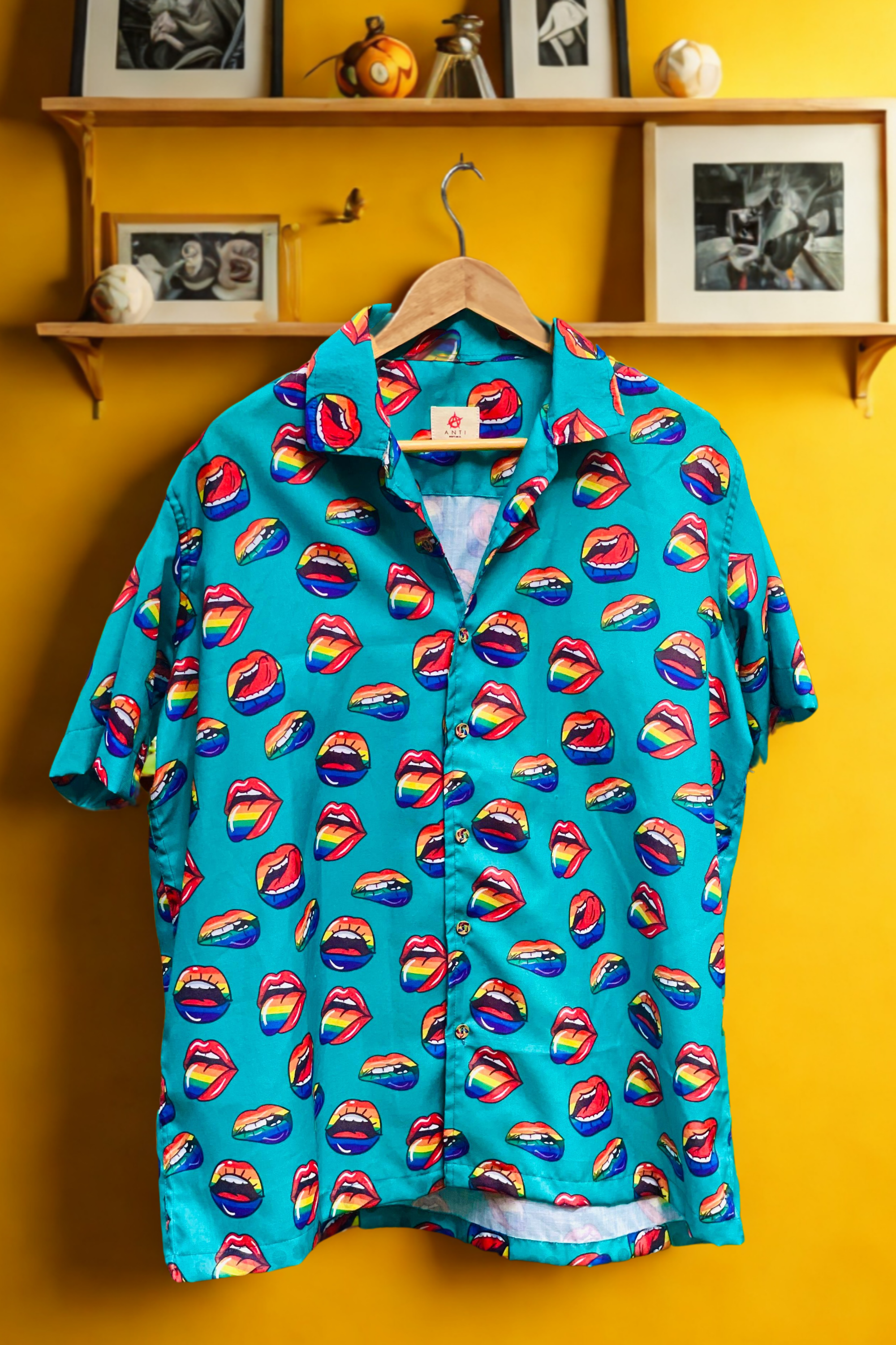 Flat-lay of a bold, unisex short-sleeve button-up shirt by ANTI, featuring a pop-art-style all-over print of red lips with rainbow-colored tongues on a bright aqua background. The vibrant, playful pattern celebrates queer expression and Pride identity. Designed with a classic collar and front placket, the shirt exudes retro streetwear energy and a fashion-fluid silhouette perfect for LGBTQIA+ visibility and self-expression.