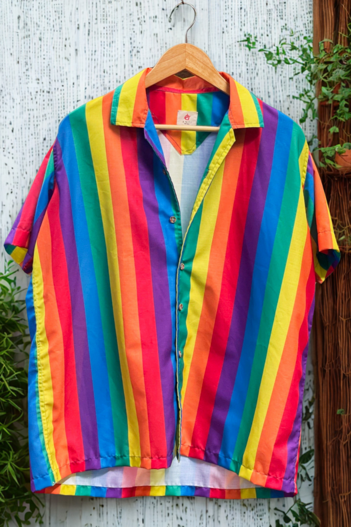 Bold rainbow-striped short-sleeve shirt on hanger against a rustic white backdrop, symbolizing queer pride and self-expression.