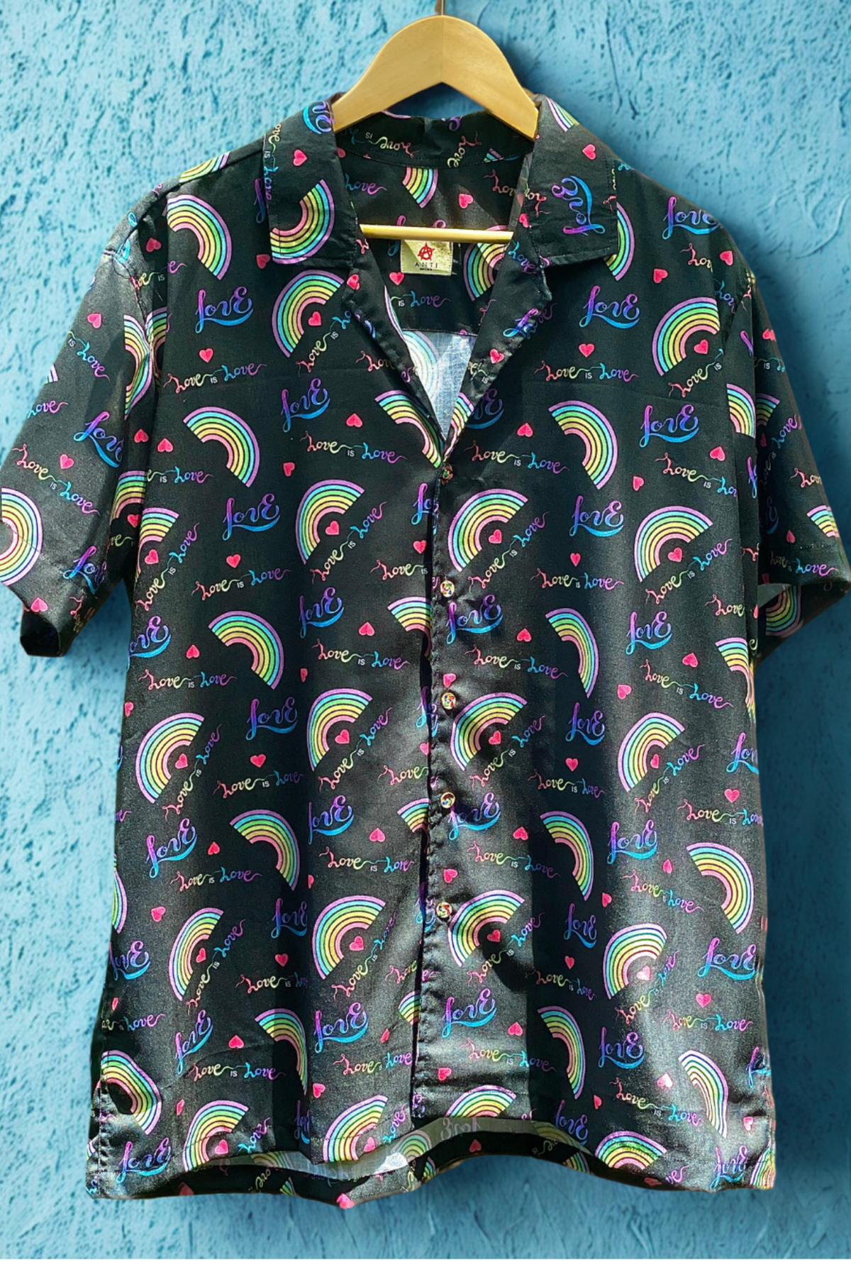 Front view of ANTI’s “Love is Love” unisex satin button-up shirt with bold rainbow hearts, multicolor text, and neon gradient graphics, designed for LGBTQIA+ visibility and inclusive streetwear style.