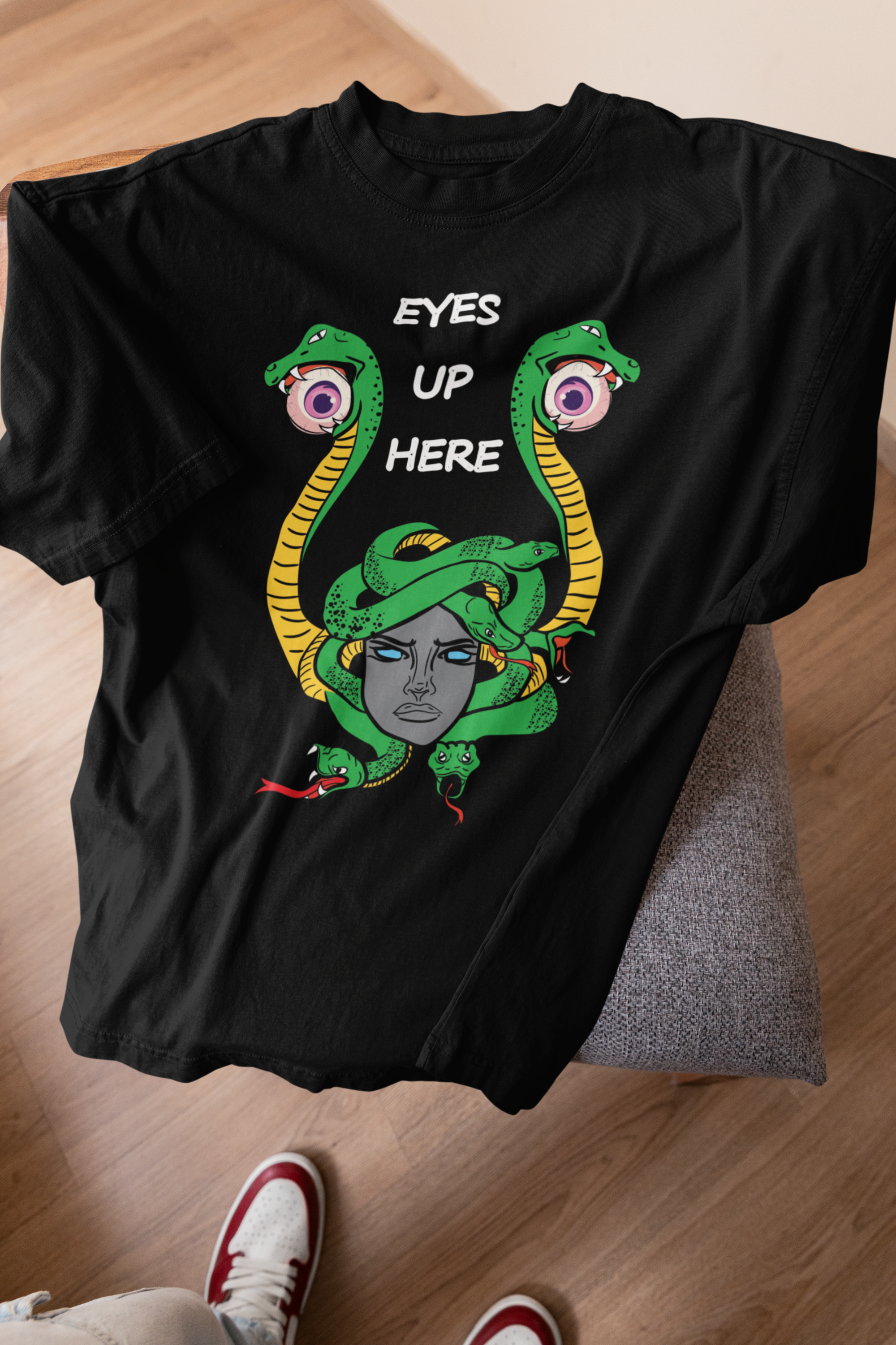 Laid-flat black t-shirt with fierce Medusa-style artwork, vibrant green and yellow snakes, and stylized “EYES UP HERE” lettering, placed on a chair with hardwood flooring and red-and-white sneakers visible below.