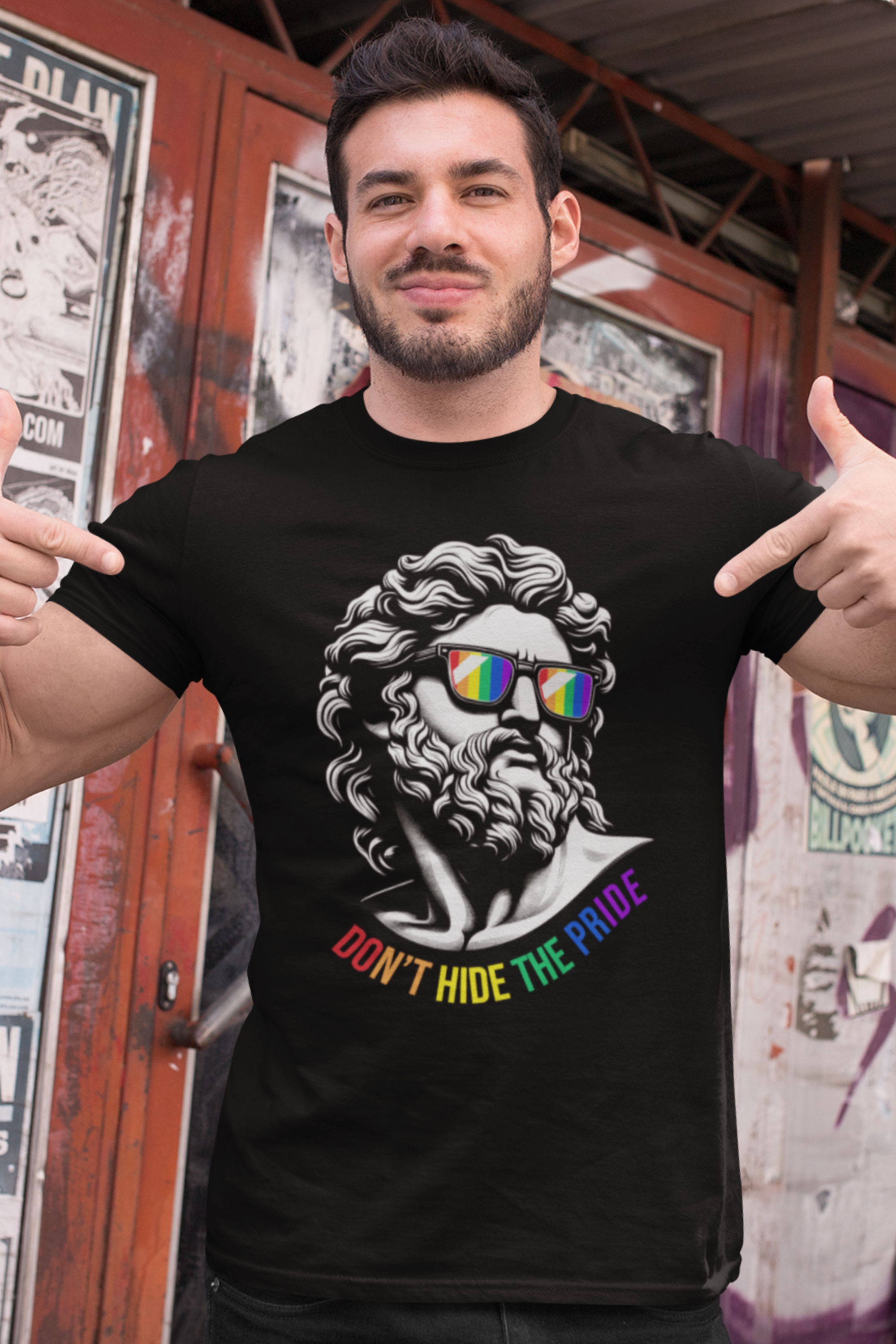 Confident man pointing to his chest while wearing ANTI’s black “Don’t Hide The Pride” t-shirt featuring a rainbow-sunglassed Greco-Roman bust, photographed outdoors in front of graffiti — celebrating LGBTQIA+ pride through bold, street-ready unisex fashion.