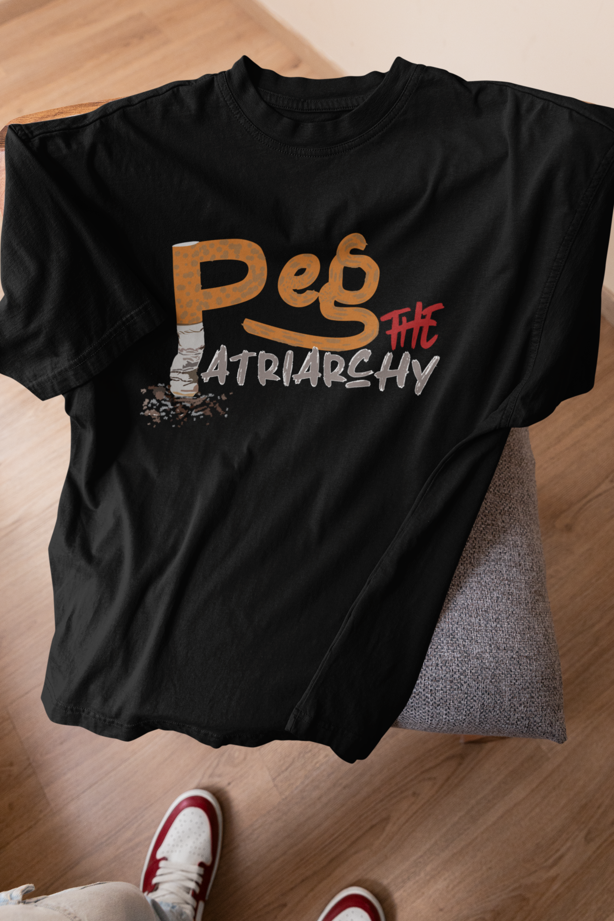 Flat lay of ANTI’s black “Peg the Patriarchy” gender-inclusive graphic t-shirt draped over a minimalist chair, captured with a sneaker-clad foot visible, offering a casual, streetwear-ready vibe for queer and ally fashion expression.

