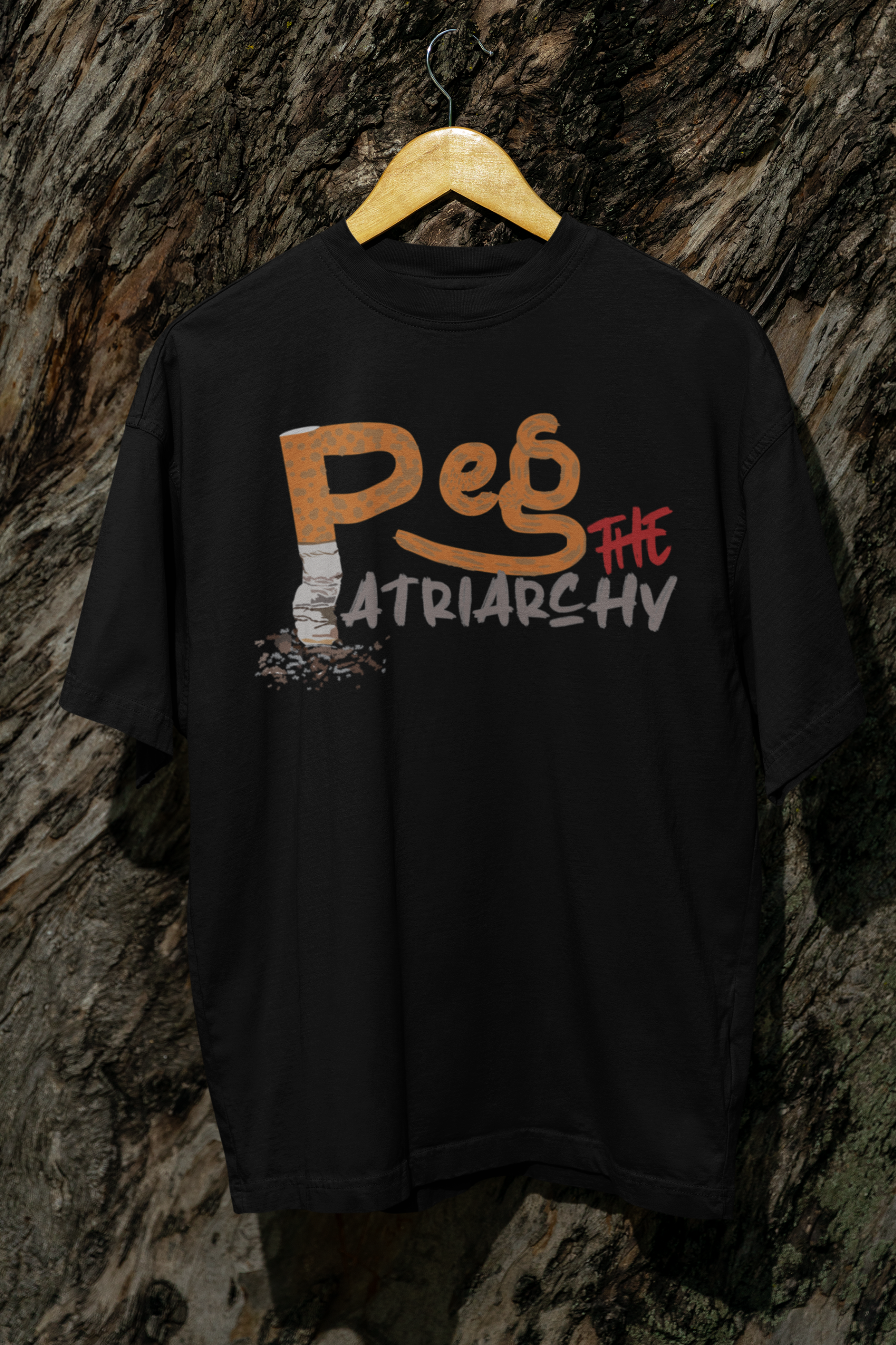 Unisex black “Peg the Patriarchy” t-shirt by ANTI, featuring a distressed cigarette butt forming the bold text, hanging against a rugged tree bark background—symbolizing resistance, defiance, and eco-aware rebellion in fashion.

