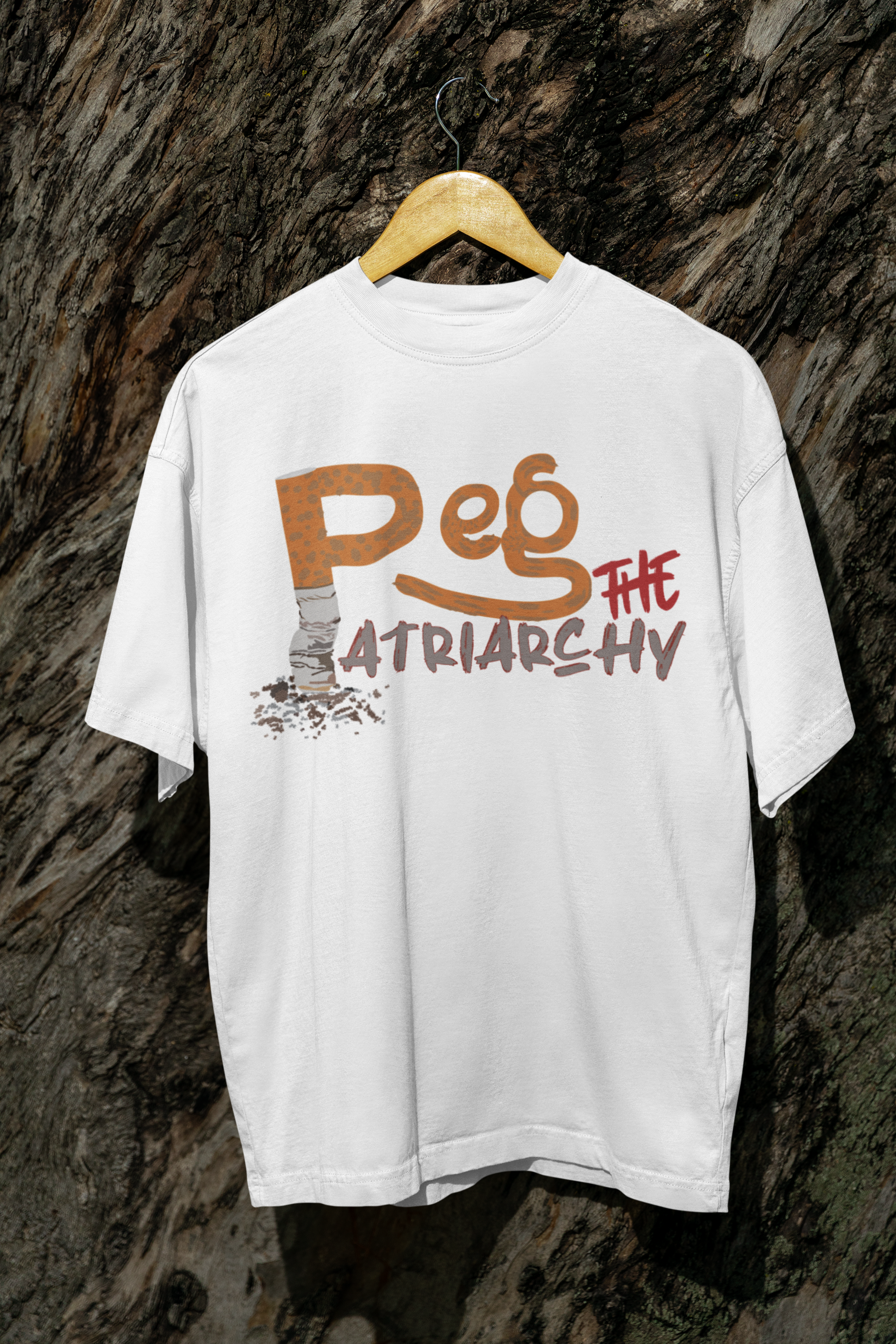 Oversized white unisex t-shirt with bold black “Peg the Patriarchy” text, hanging from a tree branch outdoors. Made from soft cotton with a relaxed, gender-fluid fit, this queer-empowered fashion piece challenges norms and celebrates inclusive expression.