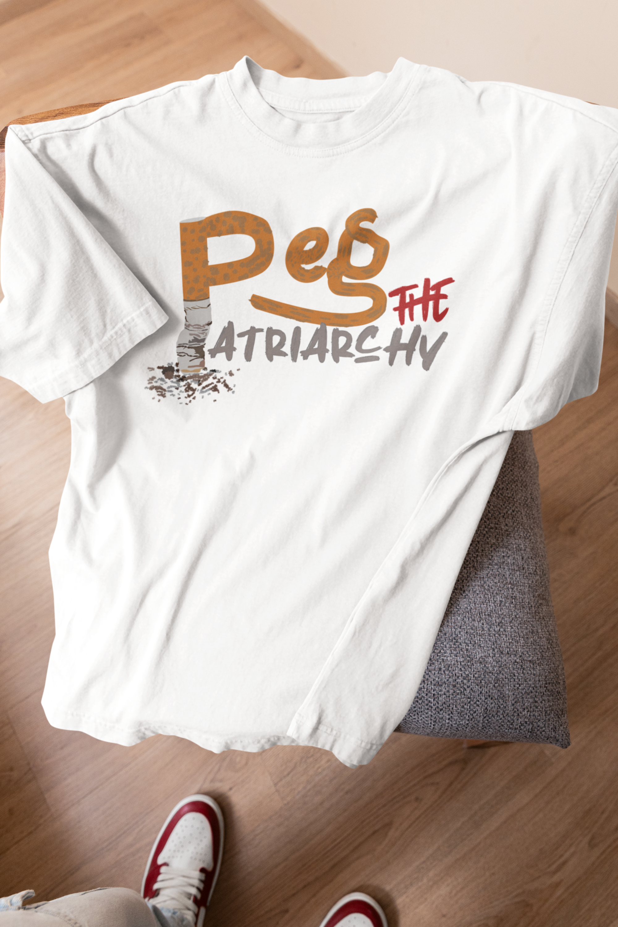 Flat lay of a white ANTI t-shirt on a chair, showcasing a vibrant and disruptive graphic reading “Peg the Patriarchy” in street-style typography with orange, red, and grey accents; a unisex, gender-fluid top challenging traditional norms.

