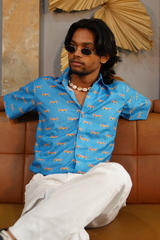 Man wearing a blue patterned shirt and sunglasses sitting on a brown couch.