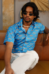 Man wearing a blue shirt with car patterns and sunglasses sitting on a brown couch.