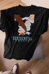 Black ANTI t-shirt laid flat on a chair with red and white sneakers visible at the bottom. The shirt features interlocked hands of different skin tones wrapped in the trans pride flag, with bold “SEXSSENTIAL CRISIS” lettering in electric blue and pink.

