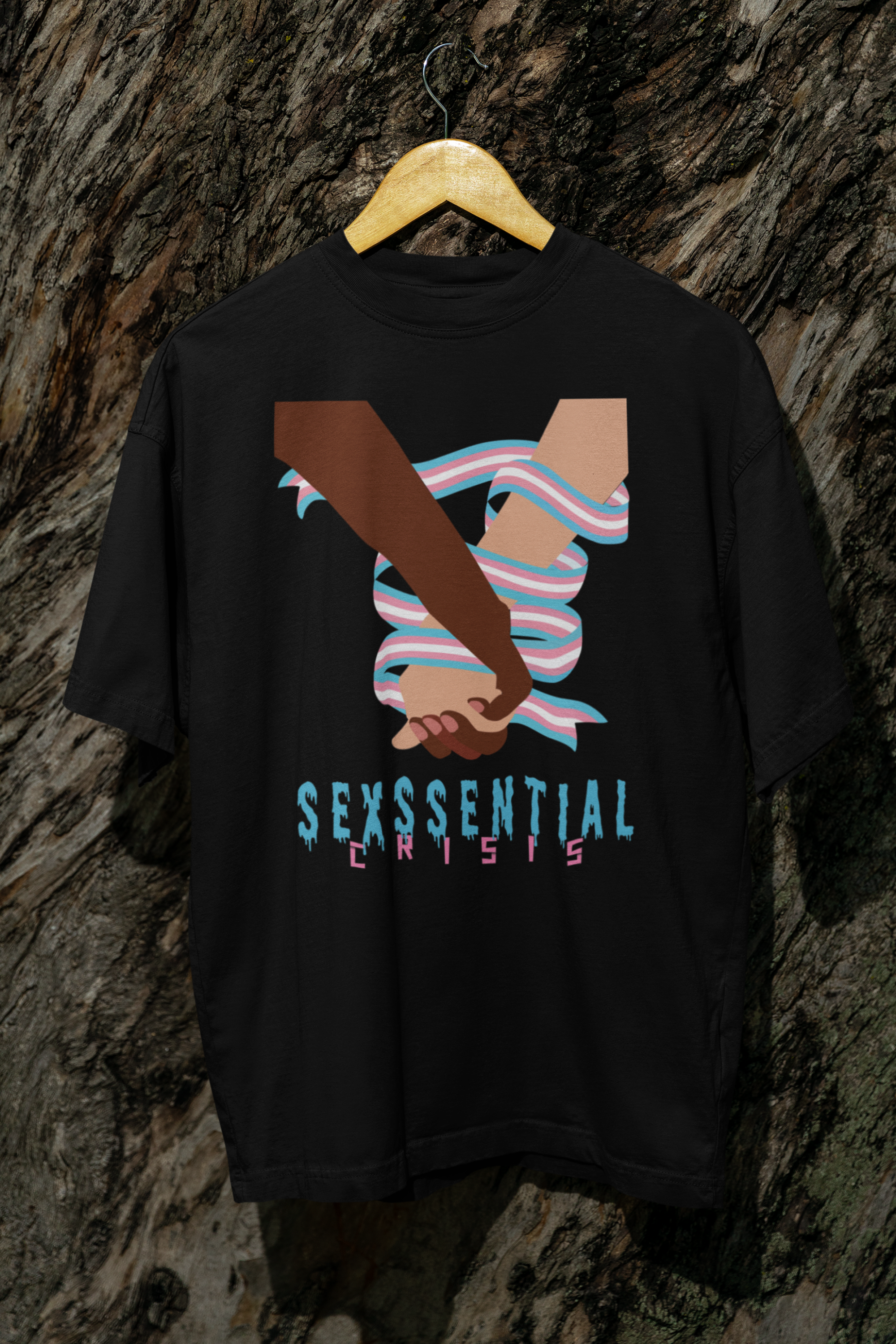 Black unisex t-shirt hanging against rugged tree bark, with a vibrant illustration of interracial hands bound by the trans pride flag, topped with the phrase “SEXSSENTIAL CRISIS” in edgy, stylized fonts. A bold tribute to queer resistance and intersectional pride.

