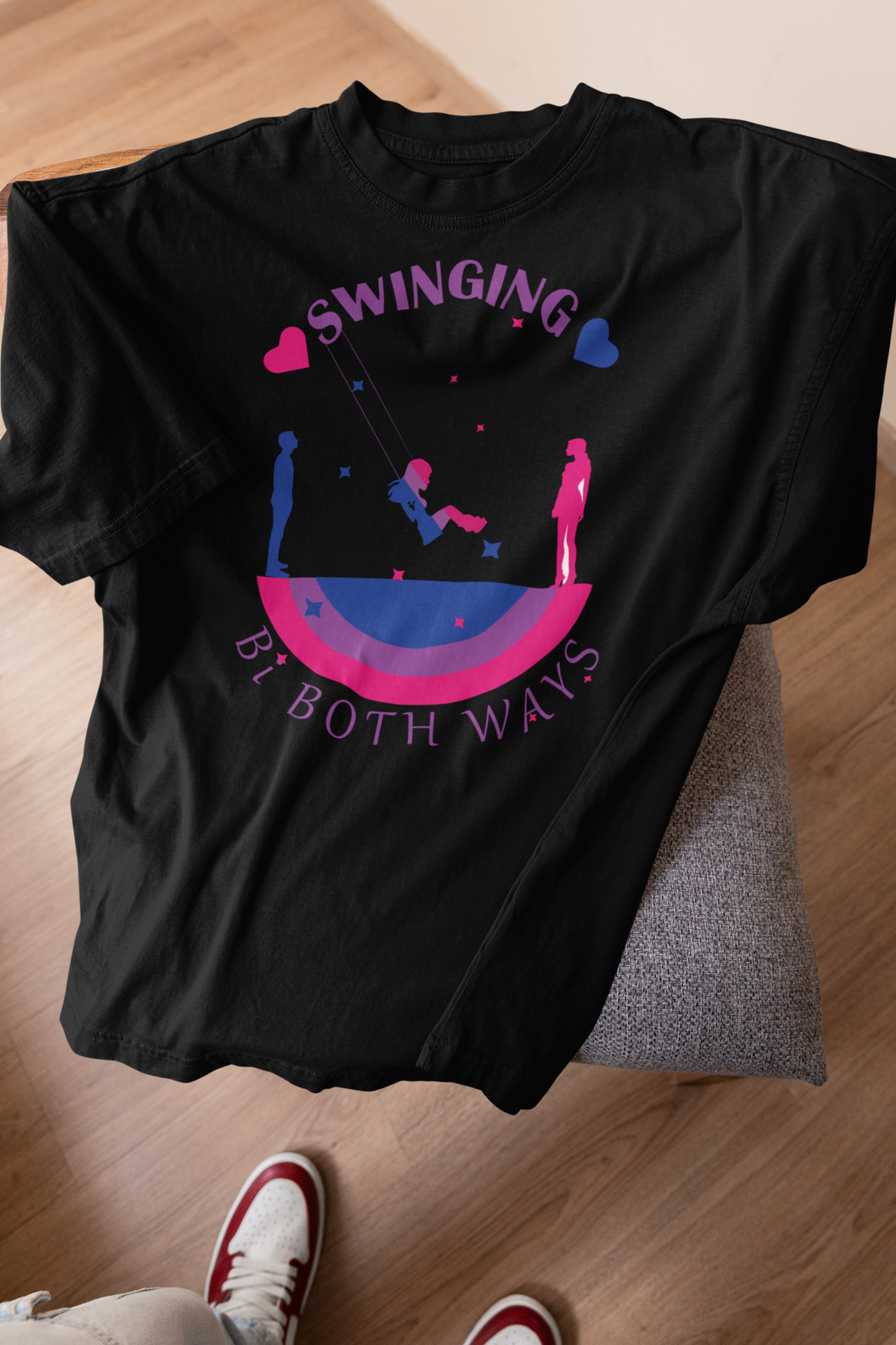 Black bisexual pride T-shirt resting on a chair, featuring a graphic with a central swinging figure, silhouettes of diverse genders, bi flag colors, and the cheeky phrase “Swinging Bi Both Ways.” Gender-fluid fashion that celebrates bi visibility and playful queer confidence.

