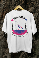 White bisexual pride graphic T-shirt displayed on a wooden hanger in front of tree bark. Design includes a swing silhouette and bi flag color arc beneath the phrase “Swinging Bi Both Ways,” symbolizing romantic fluidity with bold, inclusive flair.

