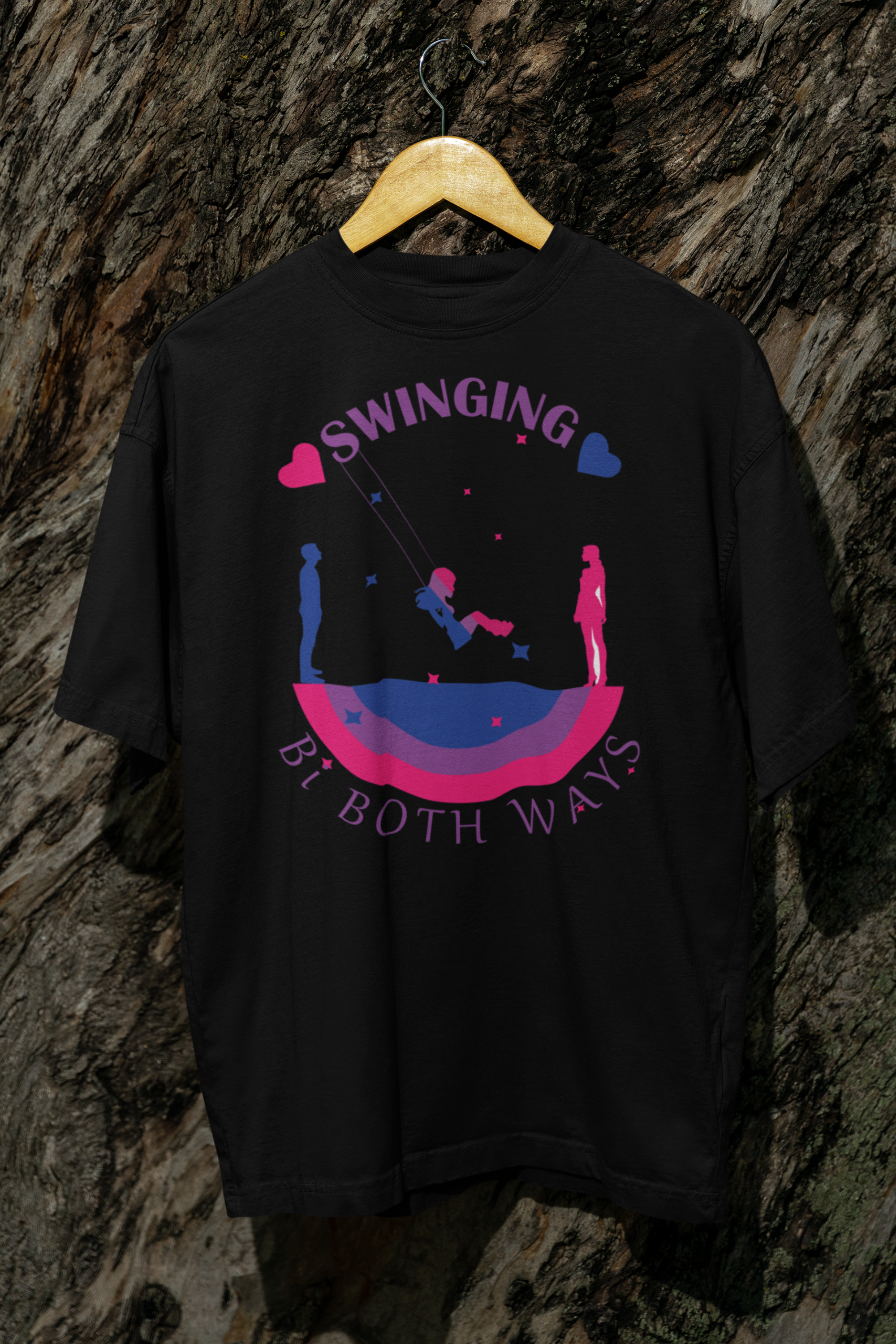 Black gender-inclusive T-shirt hung against tree bark, showing a central design with a swinging figure between male and female silhouettes over a bi flag arc. “Swinging Bi Both Ways” text celebrates bisexual identity and spectrum attraction in a bold, statement-making design.

