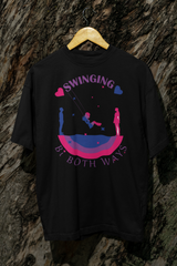 Black gender-inclusive T-shirt hung against tree bark, showing a central design with a swinging figure between male and female silhouettes over a bi flag arc. “Swinging Bi Both Ways” text celebrates bisexual identity and spectrum attraction in a bold, statement-making design.

