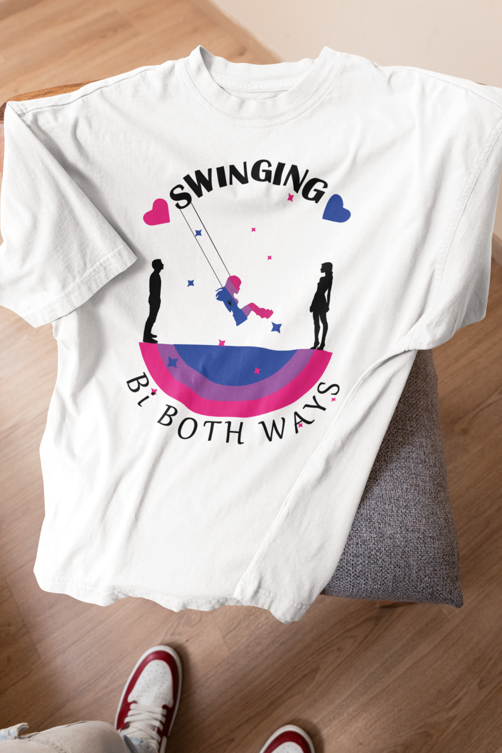 White unisex T-shirt laid on a chair, featuring a bold graphic of a person swinging between two silhouettes, framed by bi pride colors and the phrase “Swinging Bi Both Ways.” Playful and empowering design celebrates bisexual identity and fluidity in a gender-neutral cut.

