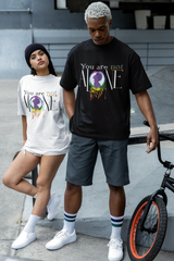 Two gender-fluid models wearing oversized ANTI t-shirts that read "You are not ALONE" with a rainbow-dripping silhouette graphic in the center. One model wears a white tee styled as a streetwear dress with white sneakers and a black beanie; the other wears a black tee with charcoal shorts and white sneakers, standing beside a BMX bike. The design highlights LGBTQIA+ pride and mental health awareness through bold typography and inclusive messaging.