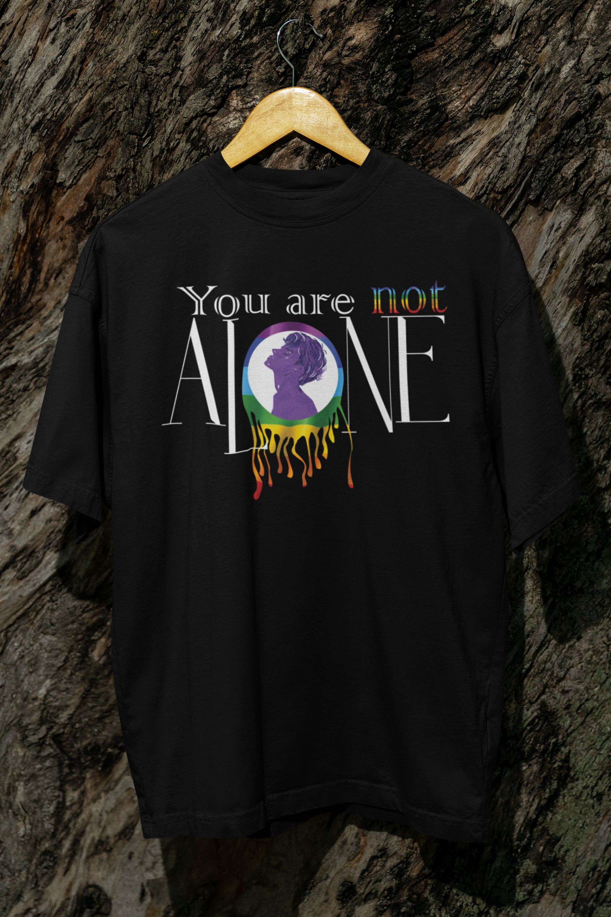 A black oversized t-shirt hanging against a rugged tree bark background, featuring the phrase “You are not ALONE” in bold white and rainbow-accented text. Centered on the shirt is a vibrant, dripping rainbow circle with the silhouette of a gender-fluid figure in purple, symbolizing LGBTQIA+ pride and mental health solidarity. The shirt is designed by ANTI, a queer-owned, gender-inclusive fashion brand.