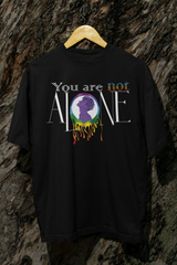 A black oversized t-shirt hanging against a rugged tree bark background, featuring the phrase “You are not ALONE” in bold white and rainbow-accented text. Centered on the shirt is a vibrant, dripping rainbow circle with the silhouette of a gender-fluid figure in purple, symbolizing LGBTQIA+ pride and mental health solidarity. The shirt is designed by ANTI, a queer-owned, gender-inclusive fashion brand.