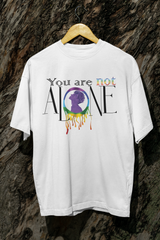 White LGBTQIA+ inclusive t-shirt displayed on a wooden hanger in front of a textured tree trunk. The bold graphic reads "You Are Not Alone," with a rainbow-infused “not” and a central figure in a rainbow ring. Dripping paint effect symbolizes emotional expression and queer pride.