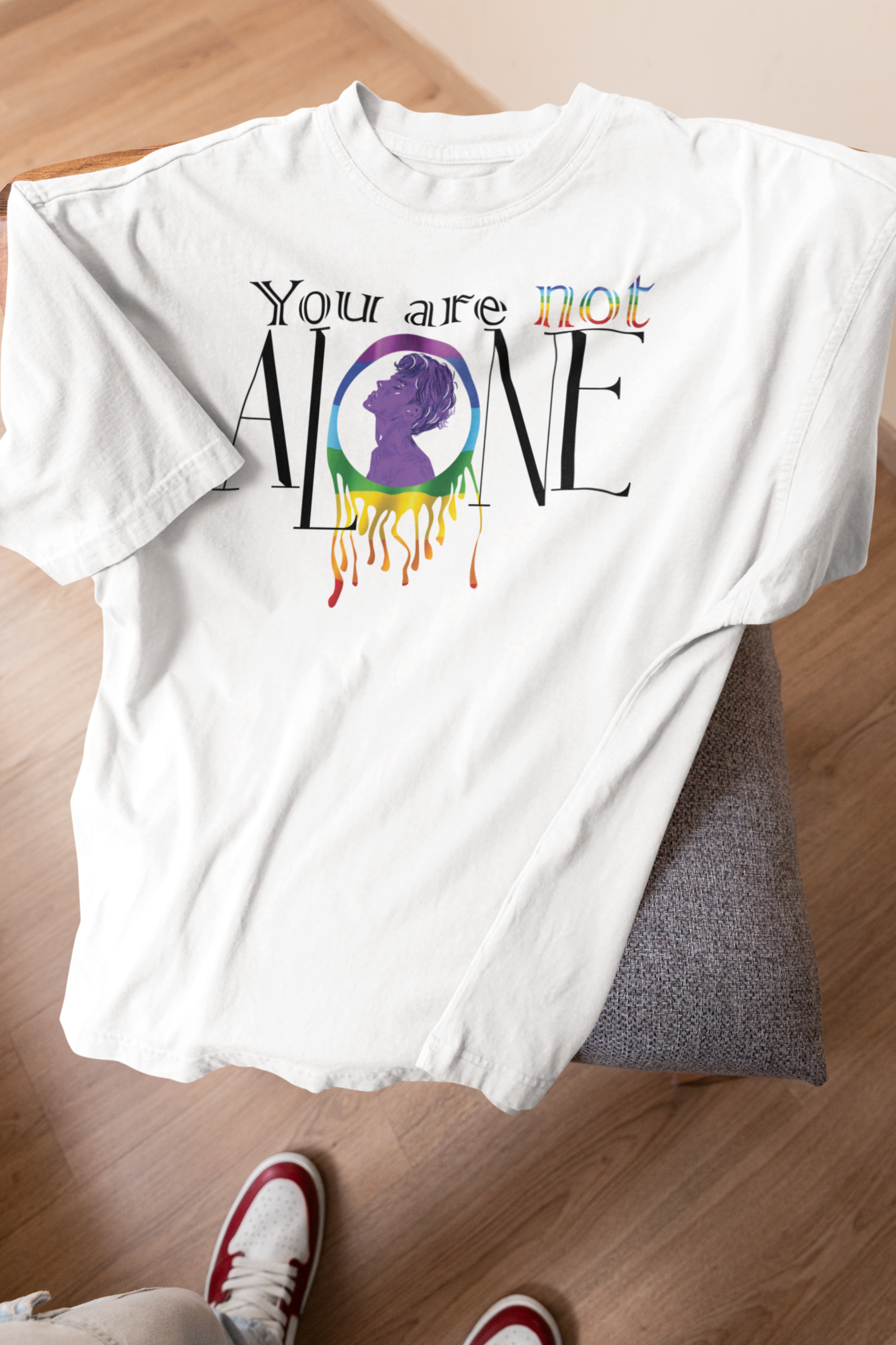 White all-gender t-shirt laid casually over a grey cushioned chair, featuring the empowering message "You Are Not Alone" in expressive typography. A central rainbow circle with a violet silhouette drips vibrant pride colors, making this shirt a strong symbol of LGBTQIA+ visibility and solidarity.