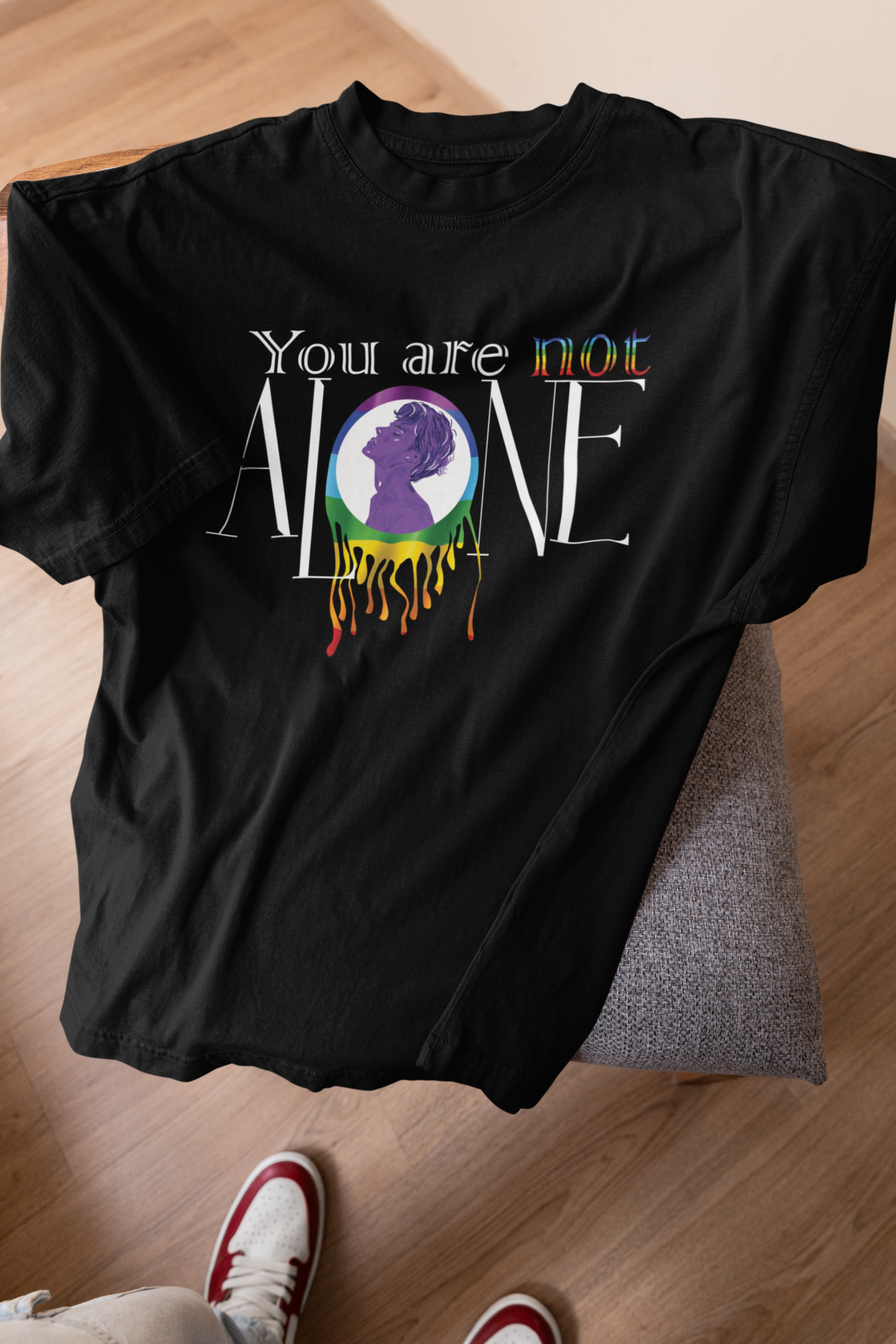 A black ANTI t-shirt laid flat on a couch, showcasing its bold “You are not ALONE” design with rainbow accents and a purple silhouette inside a melting rainbow circle. The design advocates LGBTQIA+ mental health awareness and unity. White and red sneakers and a wood floor hint at the casual, inclusive streetwear vibe of the brand’s gender-fluid collection.