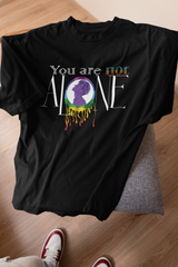 A black ANTI t-shirt laid flat on a couch, showcasing its bold “You are not ALONE” design with rainbow accents and a purple silhouette inside a melting rainbow circle. The design advocates LGBTQIA+ mental health awareness and unity. White and red sneakers and a wood floor hint at the casual, inclusive streetwear vibe of the brand’s gender-fluid collection.