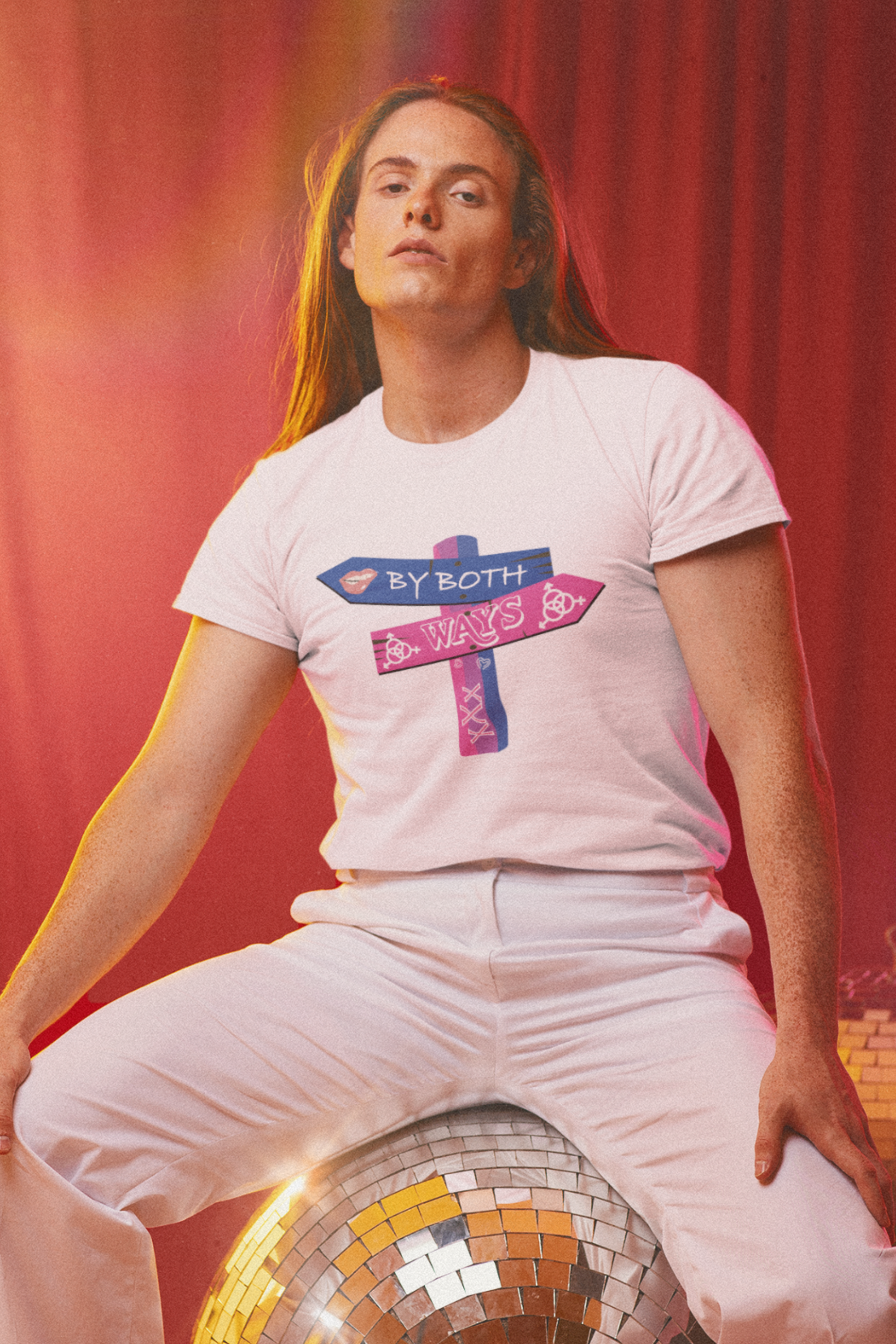 Model wearing ANTI’s “BY BOTH WAYS” white unisex t-shirt featuring a bi pride wooden arrow sign design, seated on a sparkling disco ball with a red curtain backdrop, celebrating queer identity with retro flair.

