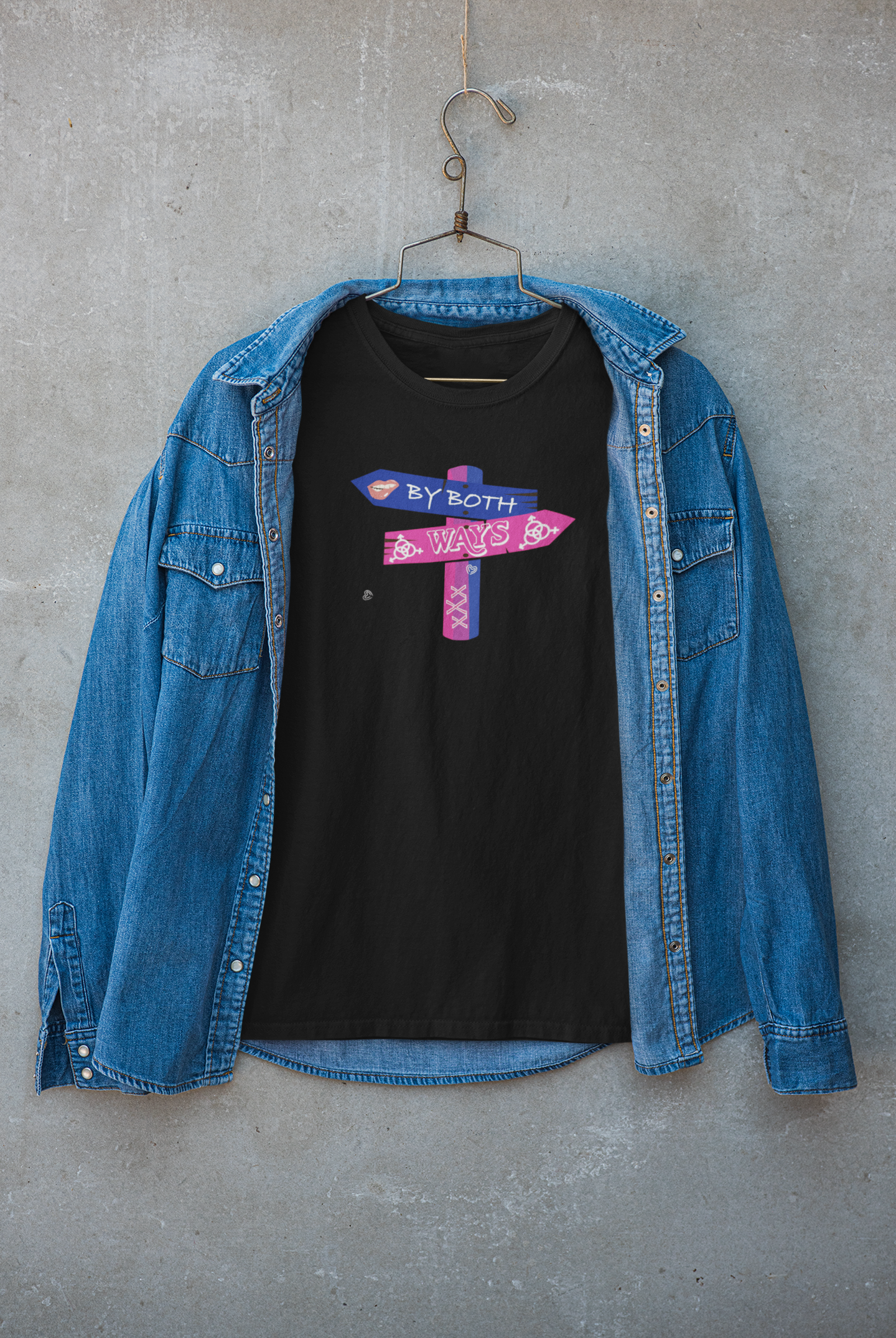 "Unisex black bisexual pride T-shirt by ANTI with 'By Both Ways' wooden sign print, styled with a denim jacket, hanging against a concrete wall – gender-inclusive, sustainable LGBTQ+ fashion."