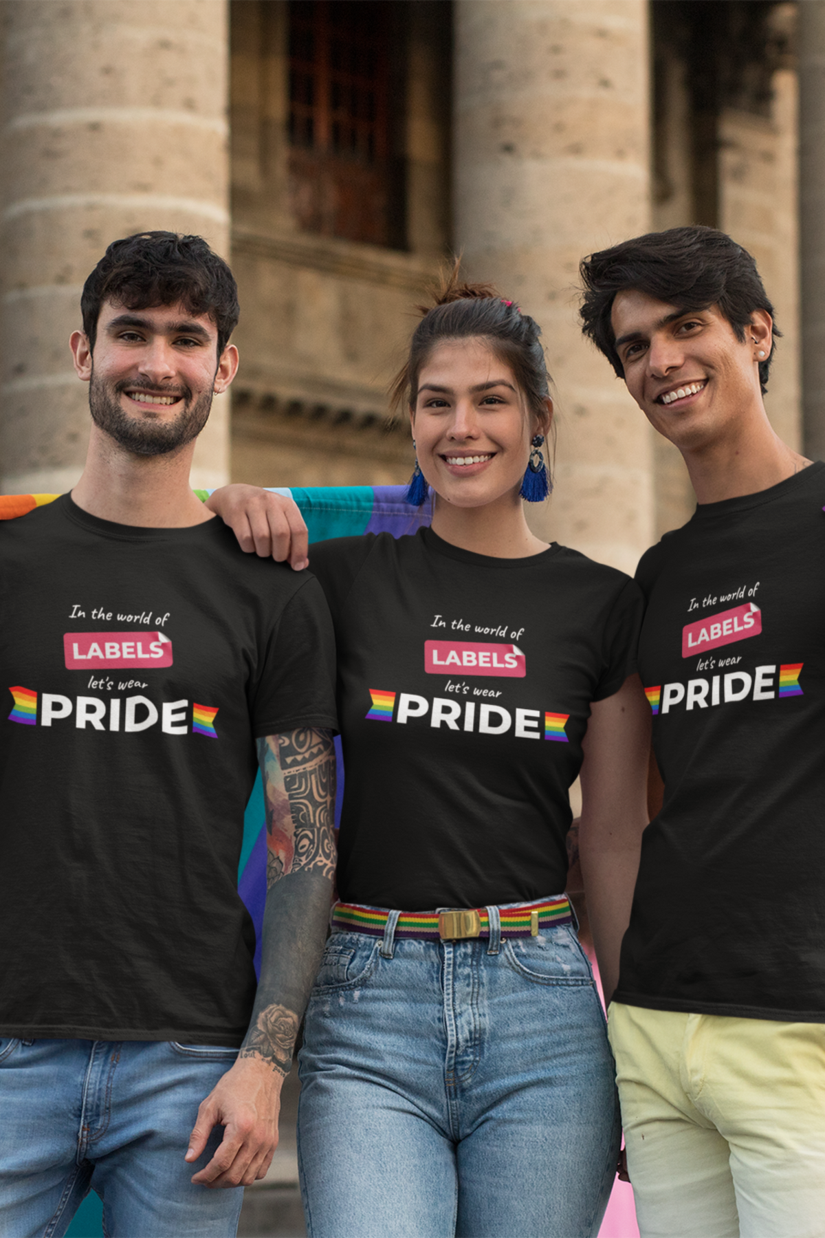 Three smiling individuals wearing black unisex “In the world of LABELS, let's wear PRIDE” t-shirts, standing confidently together in front of a stone building, showcasing inclusive LGBTQ+ fashion by ANTI.