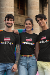 Three smiling individuals wearing black unisex “In the world of LABELS, let's wear PRIDE” t-shirts, standing confidently together in front of a stone building, showcasing inclusive LGBTQ+ fashion by ANTI.